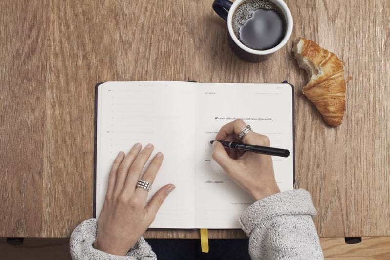 Person writing in a notebook next to coffee and a croissant